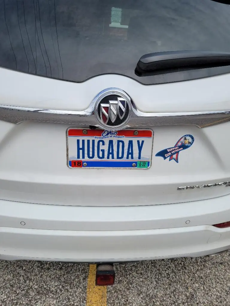 A white car with a personalized license plate reading 'HUGADAY' and a bird sticker.