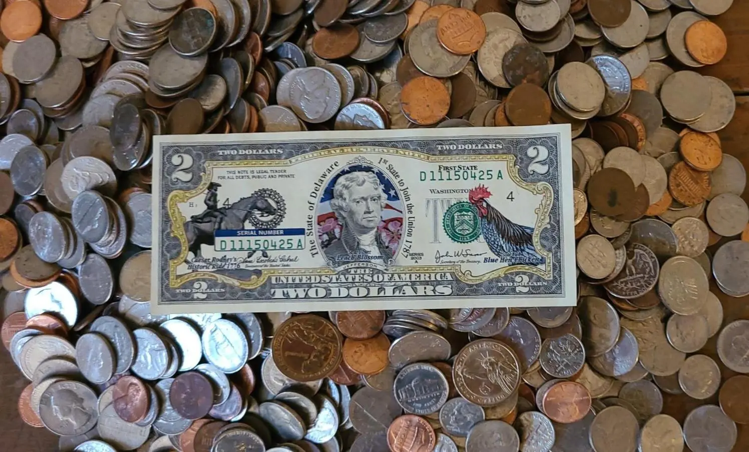 A $2 bill decorated with painted birds and a red heart, placed on a pile of coins.